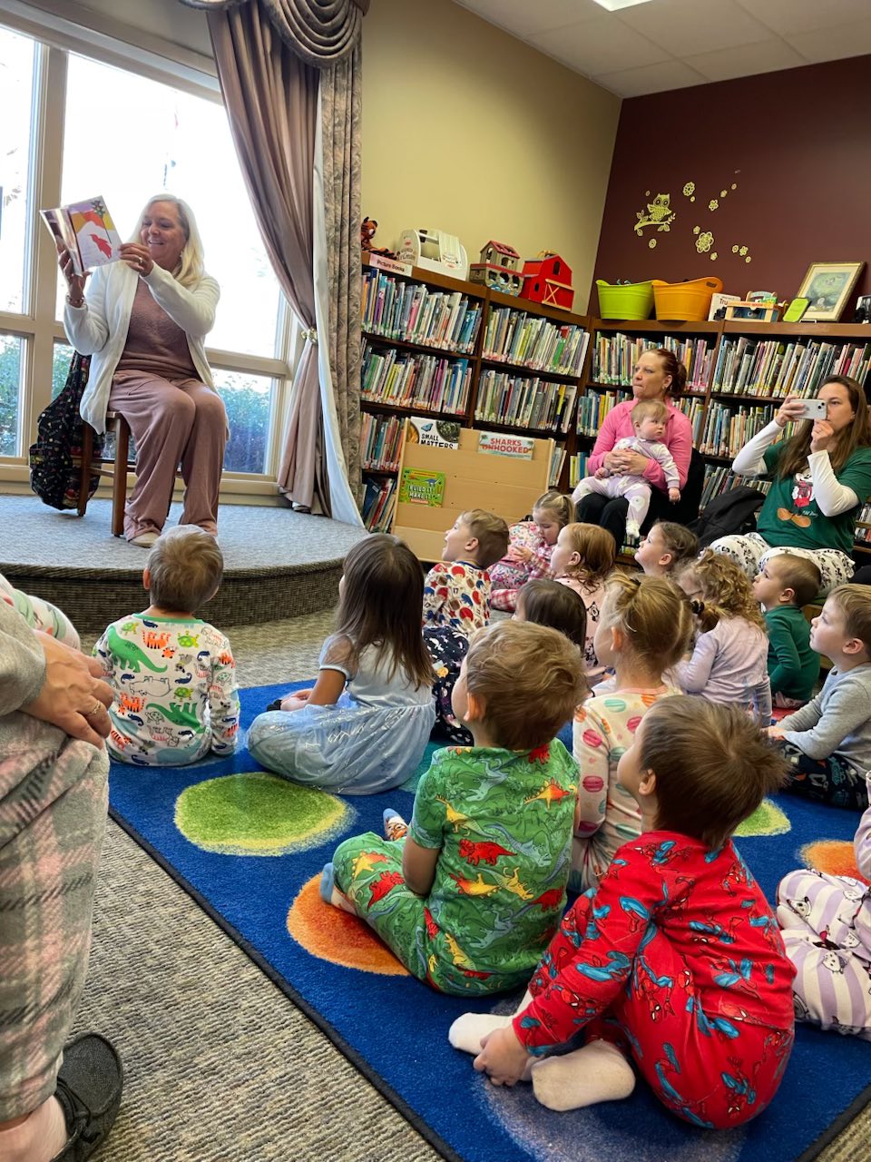 Robin reading to Rise and Shine Pre K Group A woman is reading a book to a group children age 5 and under sitting on a matt looking at her